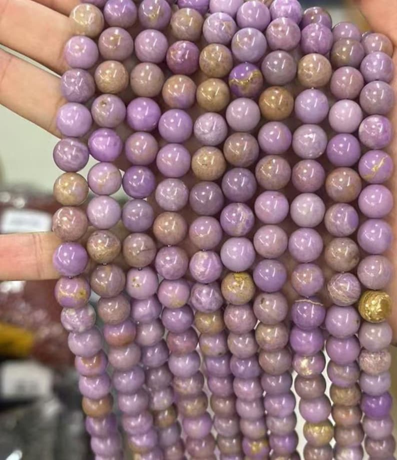 May include: Strands of round, polished beads in shades of lavender and tan. The beads are strung together, creating a textured surface. The beads are likely made of stone or glass, and are suitable for jewelry making.