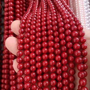 Puede incluir: Primer plano de múltiples hebras de cuentas rojas y redondas. Las cuentas son de un color rojo vibrante y brillante y parecen estar ensartadas. Las cuentas están sujetas por una mano. Las cuentas son probablemente para la fabricación de joyas.