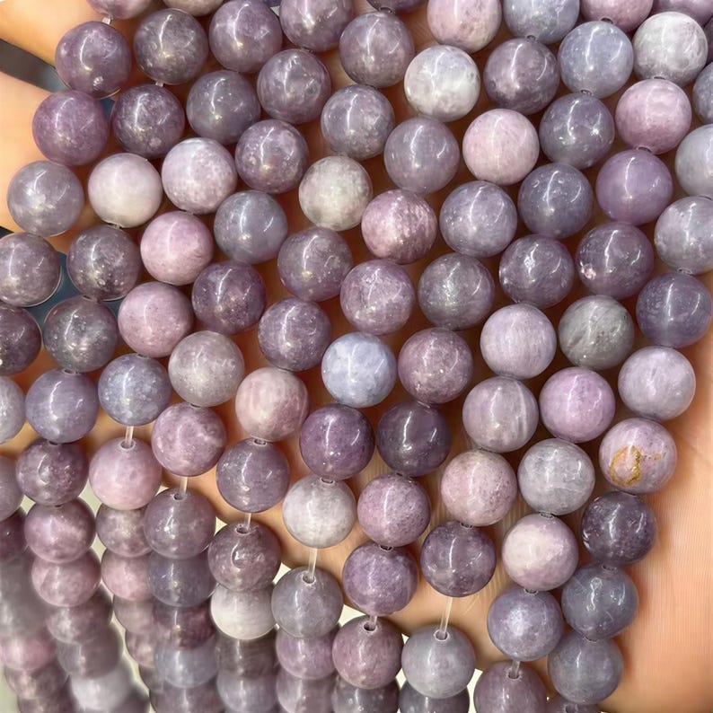 Puede incluir: Primer plano de un hilo de cuentas redondas de color lavanda. Las cuentas var&iacute;an en tonalidad, desde el p&uacute;rpura claro hasta el oscuro, algunas con un ligero brillo. Las cuentas est&aacute;n ensartadas, lo que sugiere que son para la fabricaci&oacute;n de joyas o manualidades.