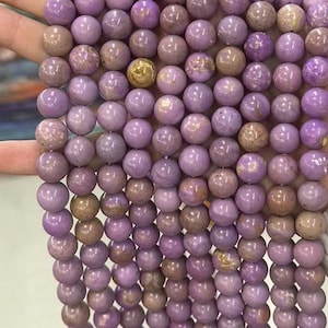 May include: Strands of round, polished beads in shades of lavender and taupe. The beads have a smooth, glossy finish and vary slightly in color. A single gold-colored bead is interspersed among the others. The beads are likely for jewelry making.