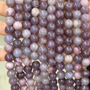 Puede incluir: Primer plano de un collar de cuentas redondas y pulidas en tonos morados, lavanda y grises. Las cuentas son lisas y parecen estar hechas de un material de piedra natural, con sutiles variaciones de color y textura.