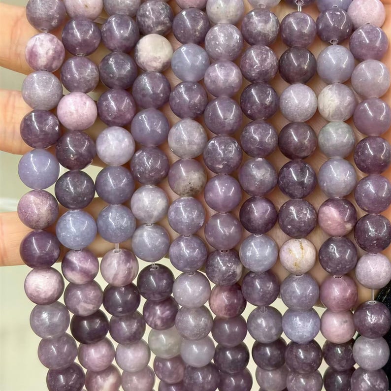 Puede incluir: Primer plano de un collar de cuentas redondas y pulidas en tonos morados, lavanda y grises. Las cuentas son lisas y parecen estar hechas de un material de piedra natural, con sutiles variaciones de color y textura.