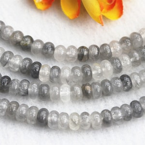 Könnte beinhalten: Eine Strähne aus grauen und klaren Quarzperlen. Die Perlen sind rund und haben eine glatte, polierte Oberfläche. Die Perlen sind auf einer durchsichtigen Gummikordel aufgefädelt.