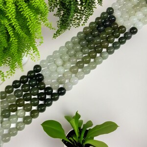 May include: A strand of round green jade beads with a gradient of color from dark green to light green. The beads are strung on a clear thread and are arranged in a line.