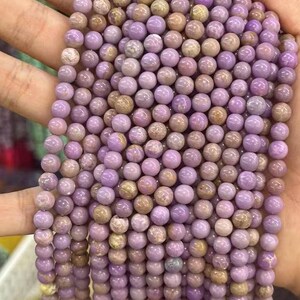 May include: Strands of round, polished beads in shades of lavender and tan. The beads are uniform in size and appear to be made of stone or a similar material. The beads are held in a hand, suggesting they are ready for use in jewelry making.