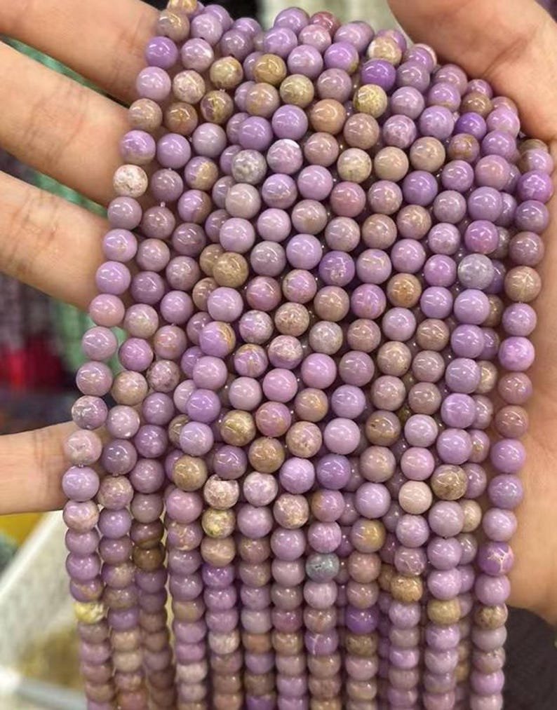 May include: Strands of round, polished beads in shades of lavender and tan. The beads are uniform in size and appear to be made of stone or a similar material. The beads are held in a hand, suggesting they are ready for use in jewelry making.