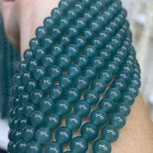 Puede incluir: Primer plano de un collar de cuentas de color verde azulado. Las cuentas son redondas y parecen lisas. Están ensartadas juntas, formando un collar o una pulsera. El color es un tono verde mar profundo. Cada cuenta mide aproximadamente 0,8 cm de diámetro.