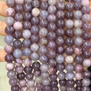 Puede incluir: Primer plano de un collar de cuentas redondas y pulidas en varios tonos de morado, lavanda y gris. Las cuentas son lisas y parecen estar hechas de piedra o un material similar. Las cuentas se sostienen en una mano.