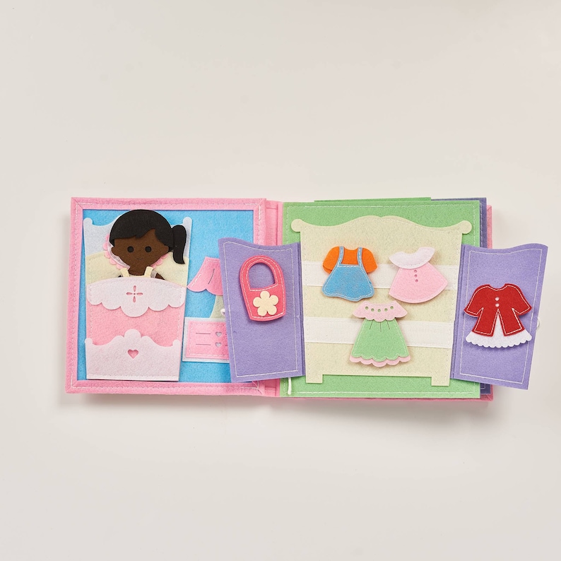 May include: A felt book with a doll in a bed on one side and a wardrobe with clothes on the other side. The doll is brown-skinned and has black hair. The wardrobe has a blue dress, a pink dress, a green dress, and a red jacket.