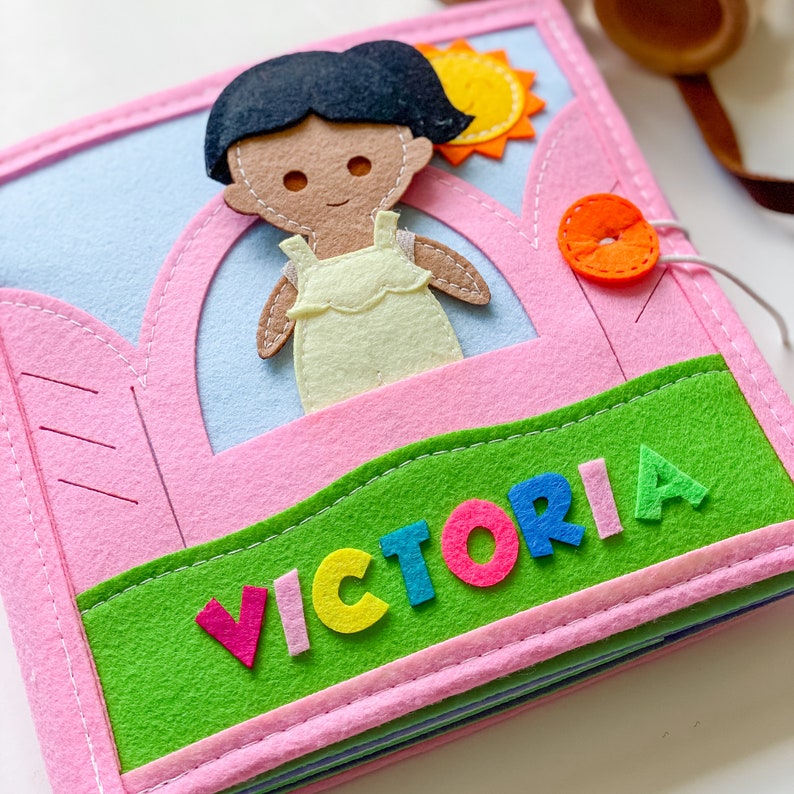 May include: A pink felt book with a green felt patch on the bottom. The patch has the name "VICTORIA" spelled out in colorful felt letters. The book has a felt doll of a girl with brown skin and black hair inside the front cover.