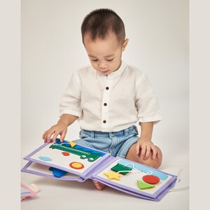 Pu&ograve; includere: Un bambino guarda un libro di attivit&agrave; in feltro con forme colorate e un autobus verde. Il libro ha una copertina lavanda e pagine con vari ritagli in feltro, tra cui un sole, una stella e forme geometriche. Il bambino indossa una camicia bianca e pantaloncini di jeans.
