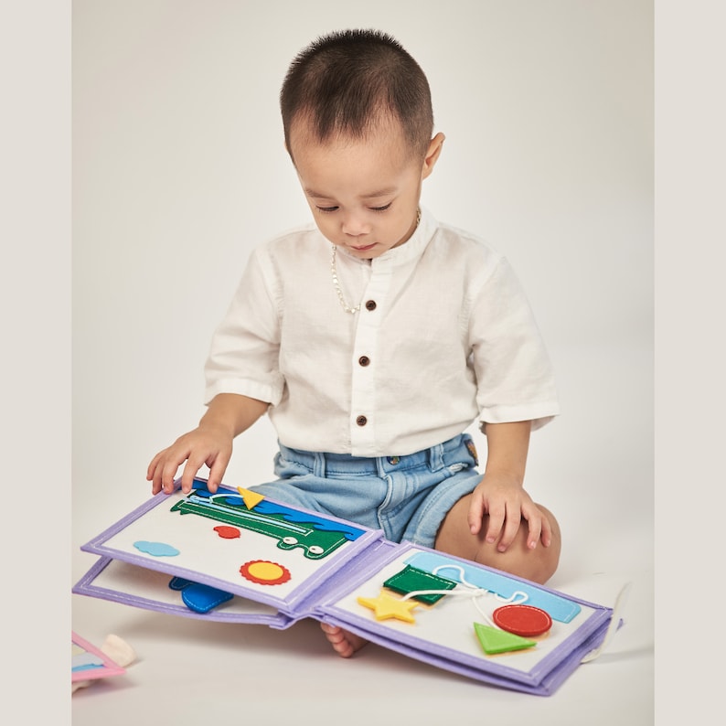 Pu&ograve; includere: Un bambino guarda un libro di attivit&agrave; in feltro con forme colorate e un autobus verde. Il libro ha una copertina lavanda e pagine con vari ritagli in feltro, tra cui un sole, una stella e forme geometriche. Il bambino indossa una camicia bianca e pantaloncini di jeans.