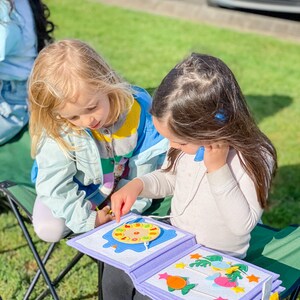 Pu&ograve; includere: Due bambini piccoli sono seduti all'aperto, concentrati su un libro di attivit&agrave; in feltro. Il libro &egrave; aperto, rivelando forme e disegni colorati in feltro. Il libro ha un bordo viola chiaro e una rilegatura bianca.