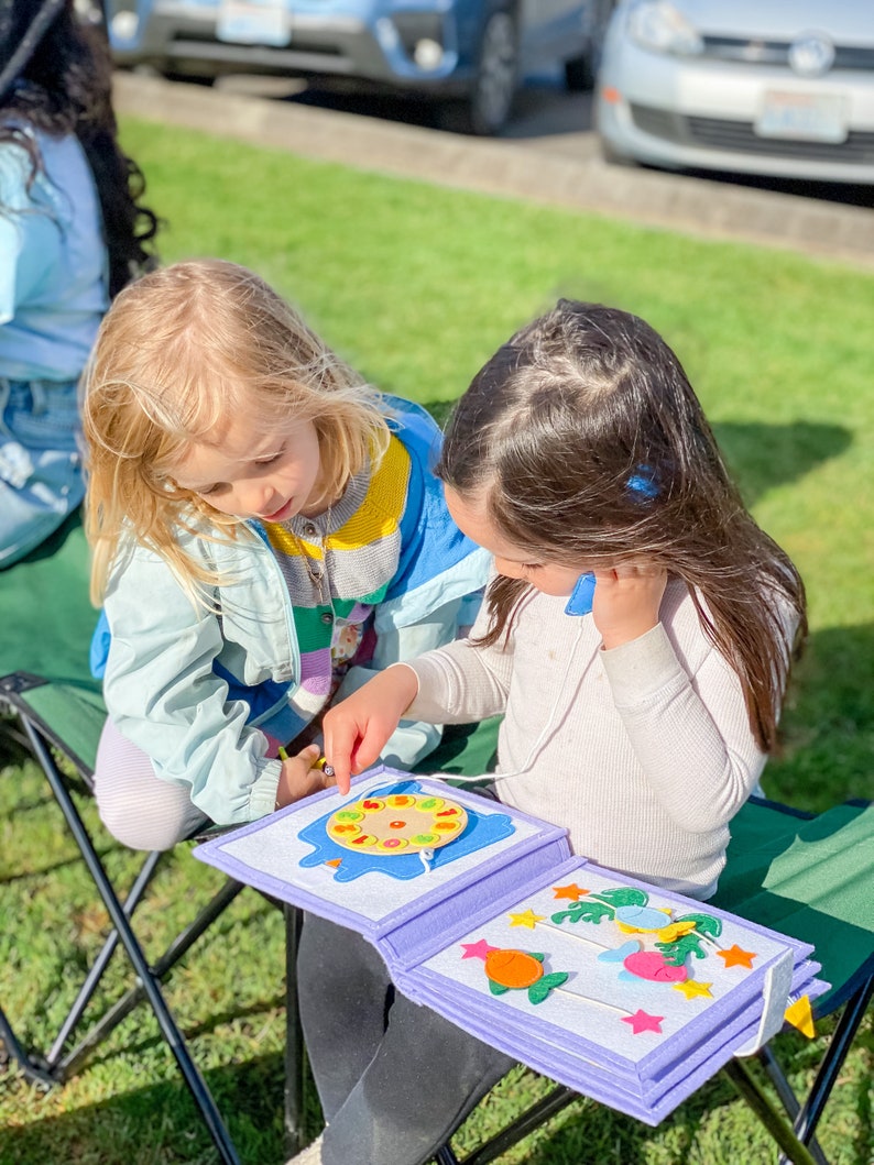 Pu&ograve; includere: Due bambini piccoli sono seduti all'aperto, concentrati su un libro di attivit&agrave; in feltro. Il libro &egrave; aperto, rivelando forme e disegni colorati in feltro. Il libro ha un bordo viola chiaro e una rilegatura bianca.