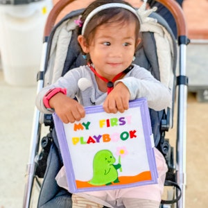 Pu&ograve; includere: Una bambina in un passeggino tiene in mano un morbido "MY FIRST PLAYBOOK". Il libro presenta un dinosauro verde che tiene un fiore. La bambina indossa una maglia grigia a maniche lunghe, una fascia e leggings fantasia.