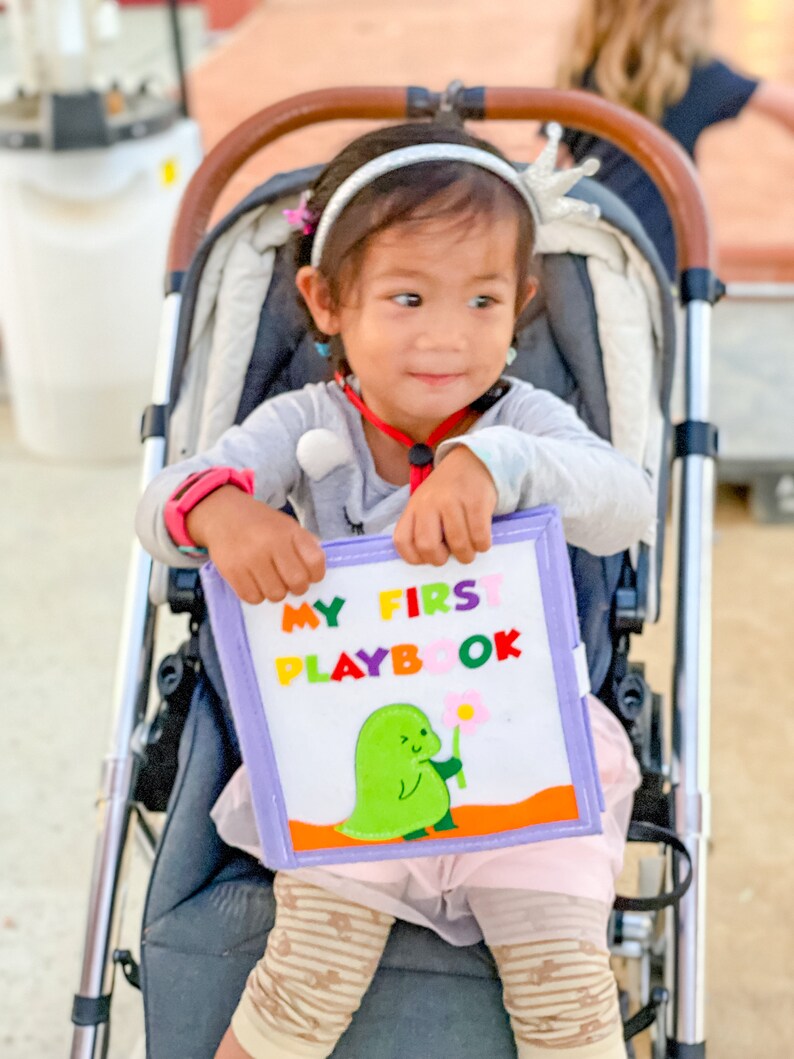 Pu&ograve; includere: Una bambina in un passeggino tiene in mano un morbido "MY FIRST PLAYBOOK". Il libro presenta un dinosauro verde che tiene un fiore. La bambina indossa una maglia grigia a maniche lunghe, una fascia e leggings fantasia.