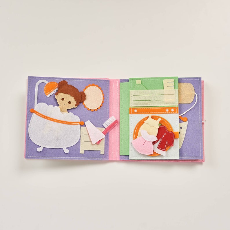 May include: A felt book with two pages. The left page shows a girl taking a bath with a shower head and bubbles. The right page shows a washing machine with a pink dress and a red shirt inside.