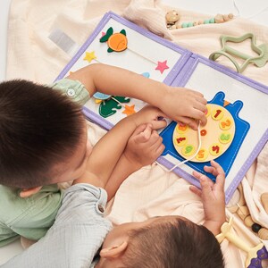 Pu&ograve; includere: Un libro interattivo in feltro con un orologio e altre forme. L'orologio ha numeri colorati e uno spago per le lancette. Il libro &egrave; bianco con un bordo viola ed &egrave; aperto, con due bambini che interagiscono con esso.