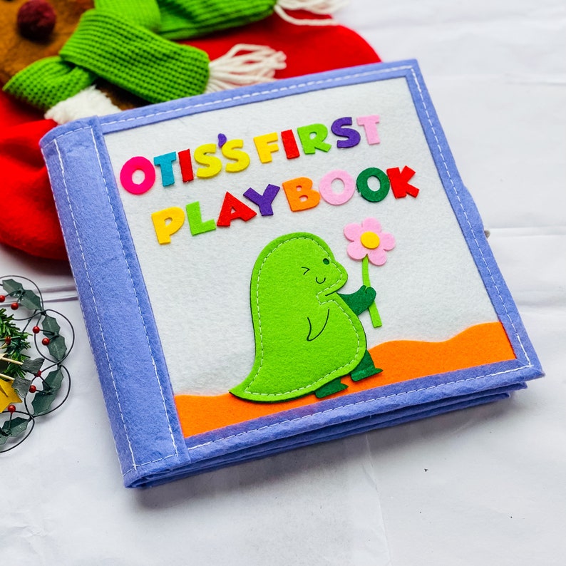 Pu&ograve; includere: Un libro gioco per bambini quadrato con un bordo azzurro. La copertina recita "OTIS'S FIRST PLAYBOOK" in lettere colorate. Un dinosauro verde tiene in mano un fiore rosa. Lo sfondo &egrave; bianco con accenti arancioni e blu.