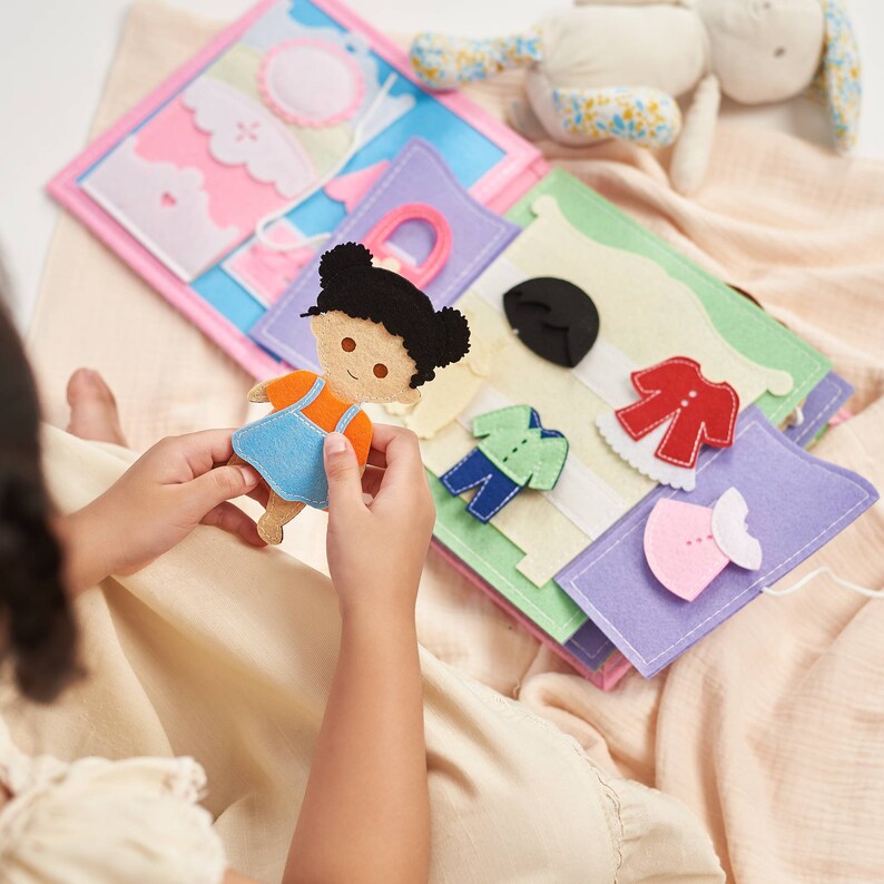 May include: A felt doll with brown hair and a blue dress is being held by a child. The doll is being dressed with different felt clothing pieces, including a red shirt, a green shirt, a blue shirt, and a pink dress. The clothing pieces are attached to a felt book with different colored pages.