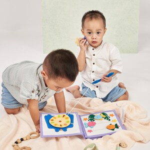 Pu&ograve; includere: Due bambini piccoli giocano con un libro di attivit&agrave; in tessuto morbido. Il libro presenta forme in feltro colorate, tra cui un orologio, pesci e stelle marine. Un bambino tiene una canna da pesca blu, mentre l'altro esamina il libro. La scena &egrave; ambientata su una morbida coperta di colore neutro.
