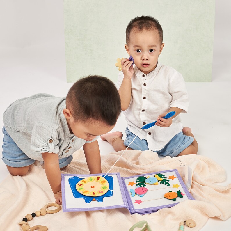 Pu&ograve; includere: Due bambini piccoli giocano con un libro di attivit&agrave; in tessuto morbido. Il libro presenta forme in feltro colorate, tra cui un orologio, pesci e stelle marine. Un bambino tiene una canna da pesca blu, mentre l'altro esamina il libro. La scena &egrave; ambientata su una morbida coperta di colore neutro.