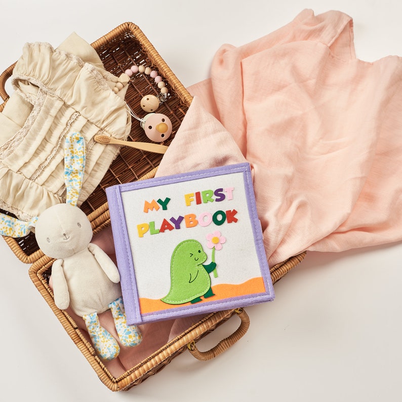 Pu&ograve; includere: Un cestino di vimini pieno di articoli per bambini, tra cui un coniglietto di peluche, un libro di stoffa con la scritta "MY FIRST PLAYBOOK", un ciuccio, uno spazzolino da denti in legno e una coperta color pesca. Il libro presenta un dinosauro verde che tiene un fiore.