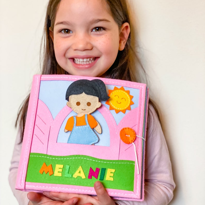 May include: A pink felt book with a girl character, a sun, and a button. The book has the name "MELANIE" on the front.