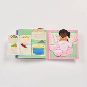 May include: A felt book with two pages. The left page shows a green felt refrigerator with a variety of felt food items inside, including a sandwich, bacon, carrots, and kiwi. The right page shows a felt table setting with a felt doll sitting at the table. The doll is wearing a blue and orange dress and has brown hair. The table setting includes a plate, a fork, a knife, a cup, and a bowl.