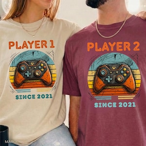 May include: Two t-shirts, one beige and one burgundy, each featuring a retro-style graphic. The graphic includes a game controller design with the text "PLAYER 1" and "PLAYER 2" above the controller, and "SINCE 2021" below.