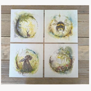 May include: Four square watercolor paintings featuring nature scenes. One shows a deer in a forest, another a bumblebee, a rabbit, and a forest scene with mushrooms. The paintings have a soft, ethereal quality.