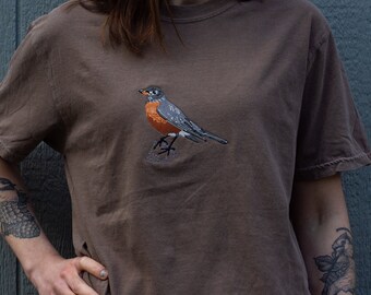 May include: A brown t-shirt with an embroidered robin on the front. The robin is gray and orange with a black beak.