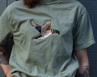 Puede incluir: Una camiseta verde salvia con un diseño de pato de madera bordado. El pato está volando con las alas extendidas y tiene la cabeza verde y el cuerpo marrón.