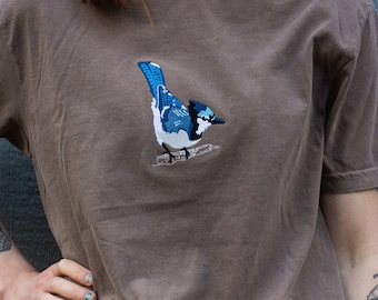 May include: A brown t-shirt with an embroidered blue jay on the front.