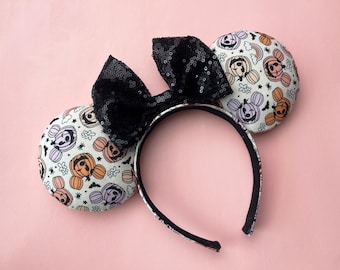 May include: Halloween-themed headband with mouse ears. The ears are patterned with smiling pumpkins in orange, purple, and peach on a white background. A large black sequined bow sits atop the headband. The headband is black and white.