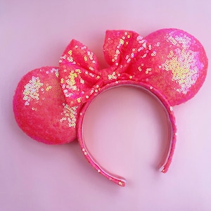 May include: A bright pink sequined headband with two round ears and a bow. The headband and ears are covered in shiny sequins, creating a sparkling effect. The bow is centered on the headband, and the ears are positioned on either side. The background is a soft pink.