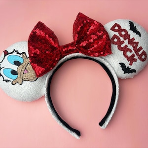 May include: A headband featuring two white sequined ears, a large red sequined bow, an embroidered Donald Duck face on the left ear, and the text DONALD DUCK with black bat silhouettes on the right ear. The headband has a black inner lining.