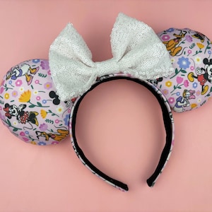 May include: A headband with two round ears and a large bow. The ears are covered in a floral pattern featuring cartoon characters. The bow is white and covered in sequins. The headband is black with a white pattern.