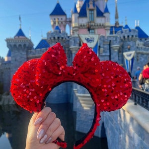 May include: Red sequined Minnie Mouse ears headband with a black headband. The headband is in front of a blue and pink castle.