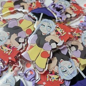 May include: A pile of anime-style acrylic keyrings. The keyrings depict two characters embracing, one with light blue hair, the other with red hair. They have purple heart-shaped clasps and are on a white surface.