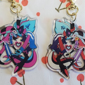 Puede incluir: Dos llaveros de acrílico transparente con una pareja de dibujos animados. La pareja es un demonio azul y rosa con cuernos y un bufón con un gorro de bufón morado y blanco. La pareja se toma de la mano y tiene corazones a su alrededor.