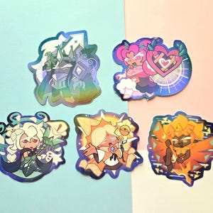 May include: Five holographic stickers featuring stylized cartoon characters. One shows a dark knight with a sword, another a pink character with a heart shield, a white-haired figure with lilies, a sun-themed character, and a fiery orange figure.
