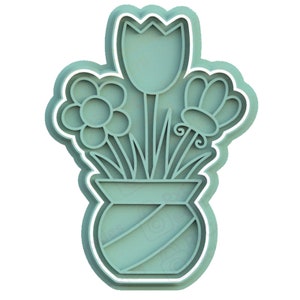 May include: A green ceramic cookie cutter in the shape of a vase with three flowers. The flowers are a tulip, a daisy, and a smaller flower with a curled stem.