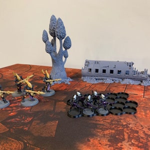 May include: A set of grey plastic miniature figures, including a group of soldiers with guns and a larger, more detailed figure of a tree. The figures are arranged on a black and red patterned surface, with a grey plastic model of a ruined building in the background.