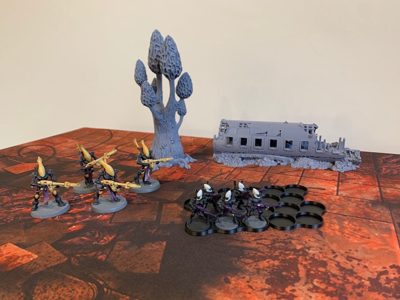 May include: A set of grey plastic miniature figures, including a group of soldiers with guns and a larger, more detailed figure of a tree. The figures are arranged on a black and red patterned surface, with a grey plastic model of a ruined building in the background.