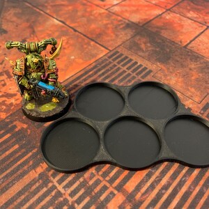 May include: Black plastic miniature painting tray with six circular compartments. The tray is designed to hold miniature figures for painting.