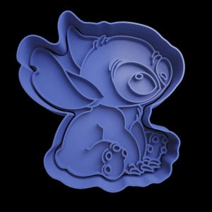 May include: A blue plastic cookie cutter depicting the cartoon character Stitch. The cutter shows Stitch in a seated position, with his arms and legs drawn in. The cutter is set against a black backdrop.
