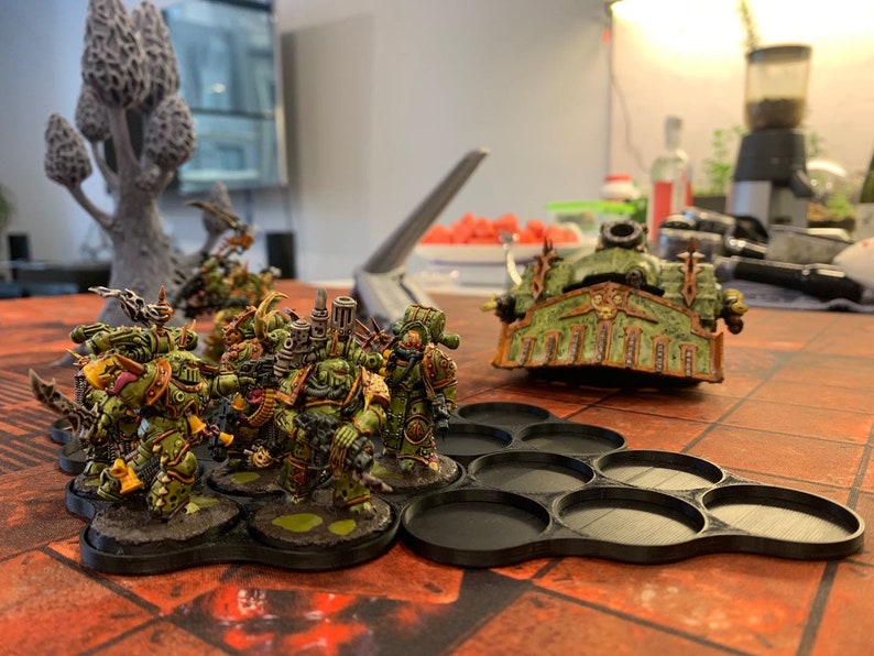May include: A group of green and yellow painted miniature figures stand on black circular bases. The figures are arranged in a line and are facing the right side of the image. A larger green and brown painted miniature figure is in the background. The figures are on a black and red patterned surface.
