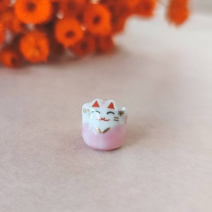 Puede incluir: Pequeña figura de cerámica Maneki Neko. El gato de la suerte es blanco y rosa con detalles dorados, una cara sonriente y orejas rojas. Es una versión en miniatura del tradicional gato de la suerte japonés.