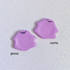 Pode incluir: Dois pingentes em forma de gota de cor lavanda com um pequeno detalhe de linha preta. Um pingente tem um acabamento brilhante e o outro tem um acabamento fosco. A palavra "glossy" est&aacute; escrita abaixo do pingente brilhante e "matte" est&aacute; escrita abaixo do pingente fosco.